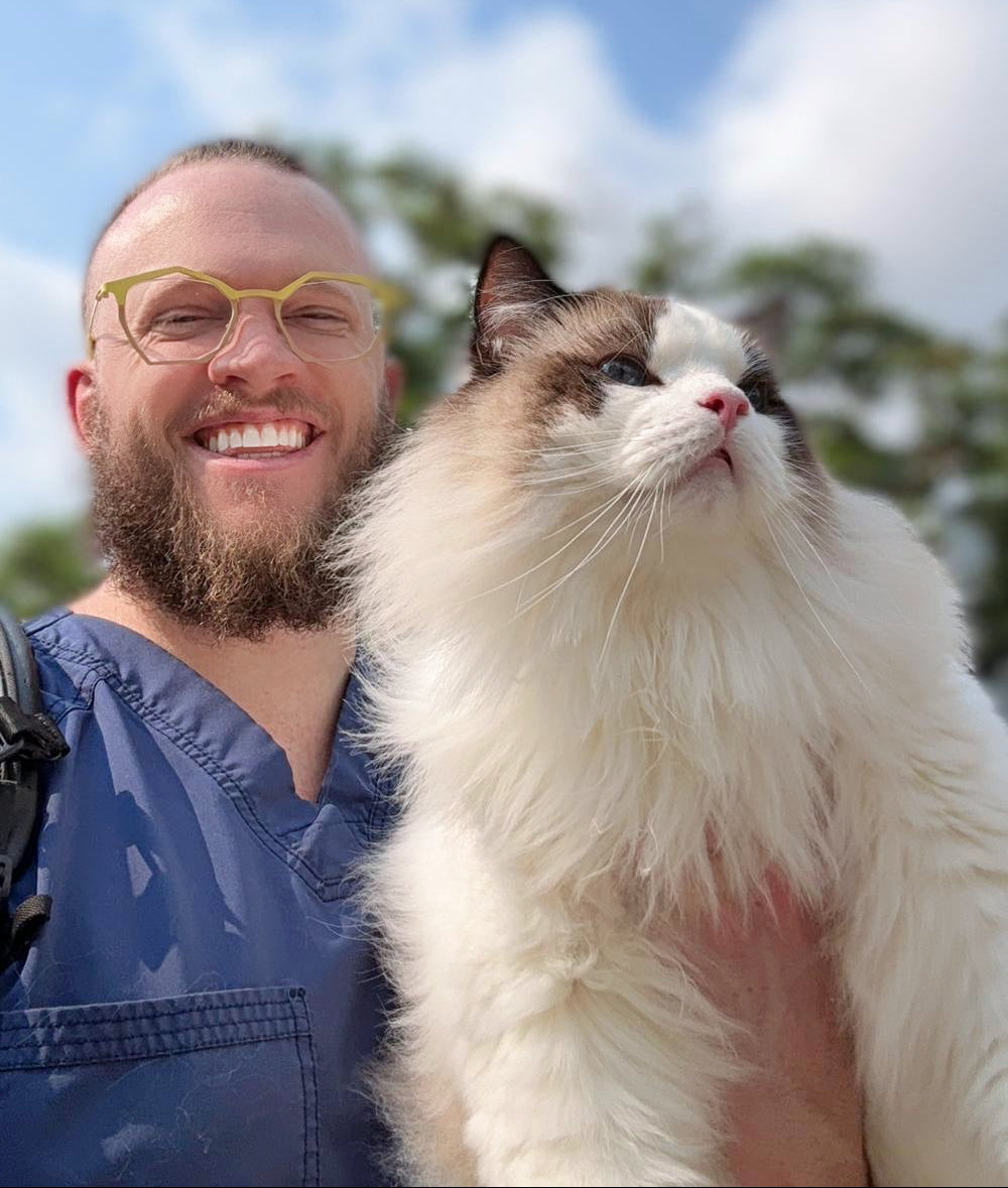 Man holding a fluffy white and brown cat against a blue sky with clouds